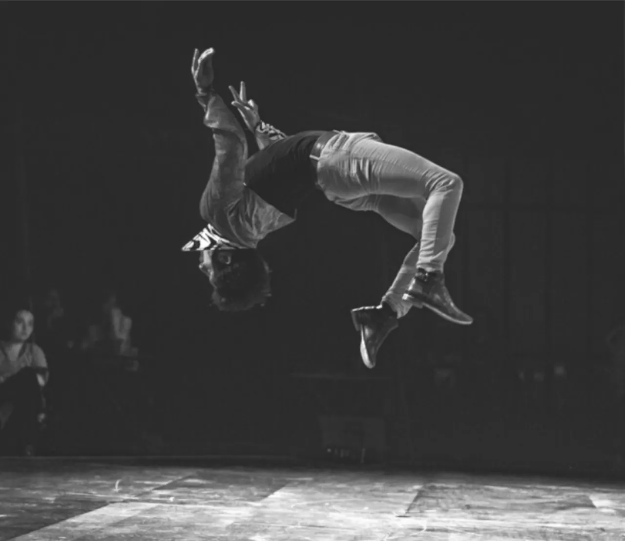 Un acrobate en plein salto arrière, tête en bas au-dessus du plateau, devant des spectateurs dans la pénombre