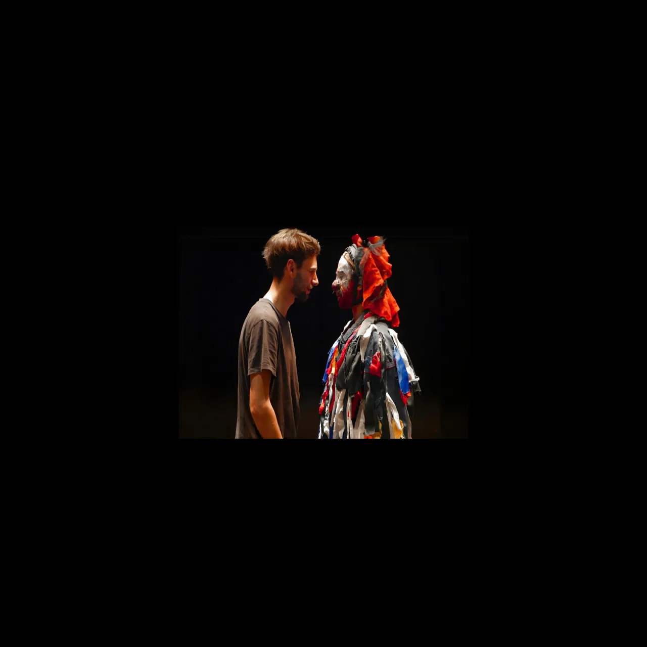 Face à face sur scène entre un comédien en t-shirt et un personnage costumé de lambeaux de tissus colorés, coiffé d'un foulard orange, le visage maquillé