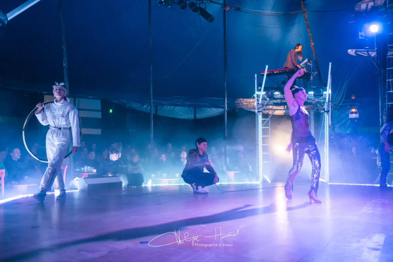 Numéro de cabaret sous chapiteau dans une lumière bleue et violette : une artiste tient un cerceau, un danseur en pantalon à paillettes lève le bras, un musicien joue en surplomb