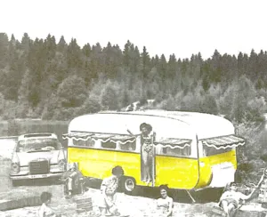 Photographie d'archive retravaillée en jaune vif : une caravane des années 1960 installée en lisière de forêt, une voiture ancienne à côté, quelques personnes autour