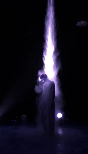 Sur un plateau plongé dans le noir, une silhouette en longue robe blanche baisse la tête sous une colonne de matière qui tombe des cintres, éclairée d'un violet intense