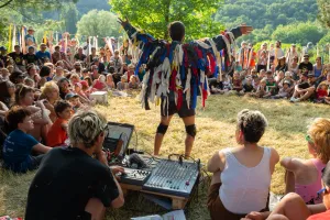 Spectacle en plein air : un artiste bras écartés, vêtu d'un costume de lanières de tissu bleues, rouges et blanches, devant un public assis dans l'herbe ; au premier plan, la table de mixage du musicien