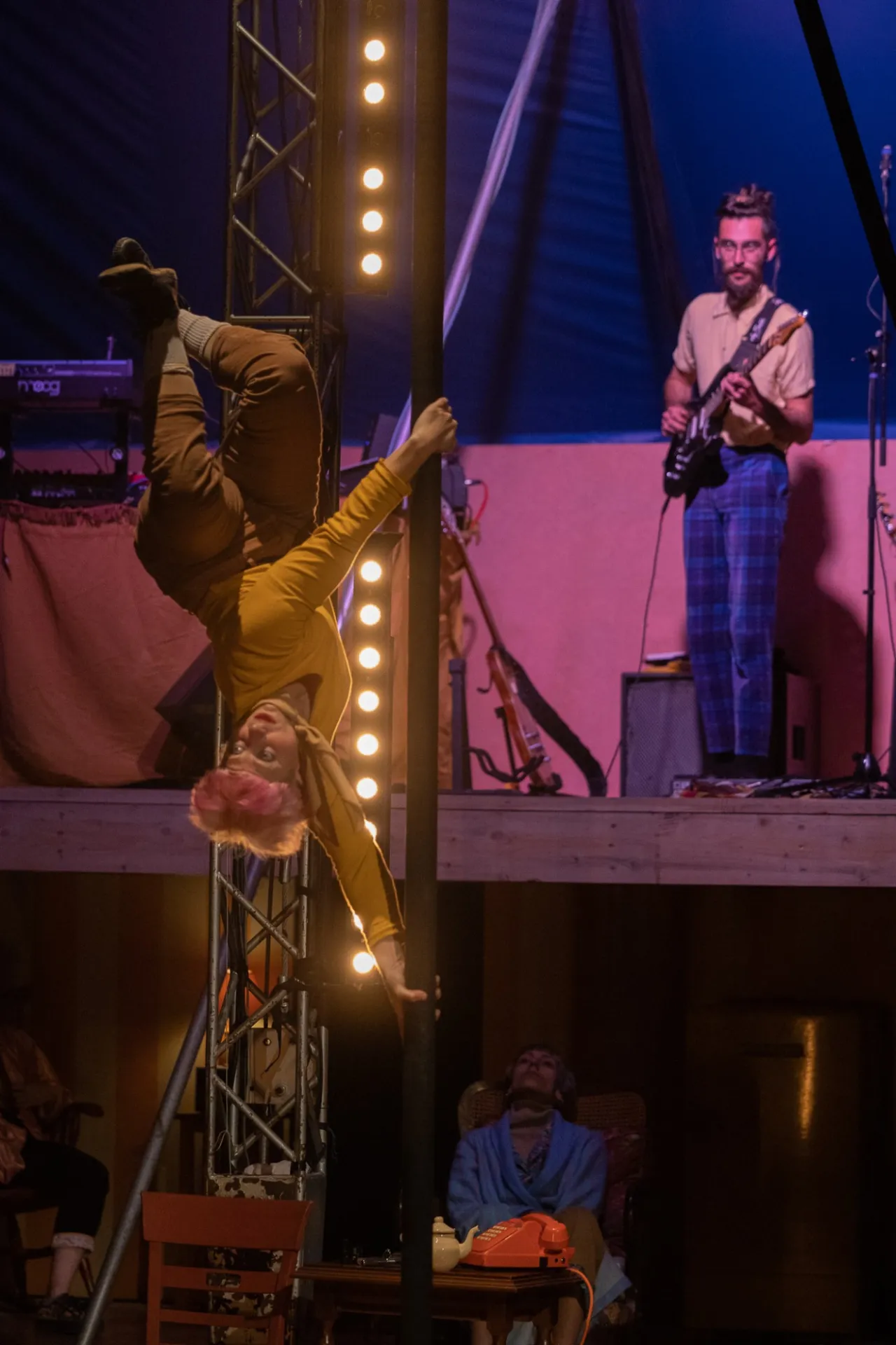 Sur la piste du Super Cabaret, une acrobate en tenue jaune se tient tête en bas le long d'un mât, pendant qu'un guitariste joue sur une estrade rose